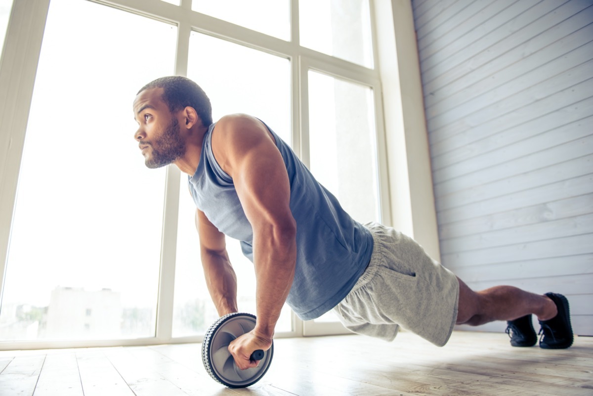 American sportsman is doing ab wheel rollout exercise and looking forward while working out at home