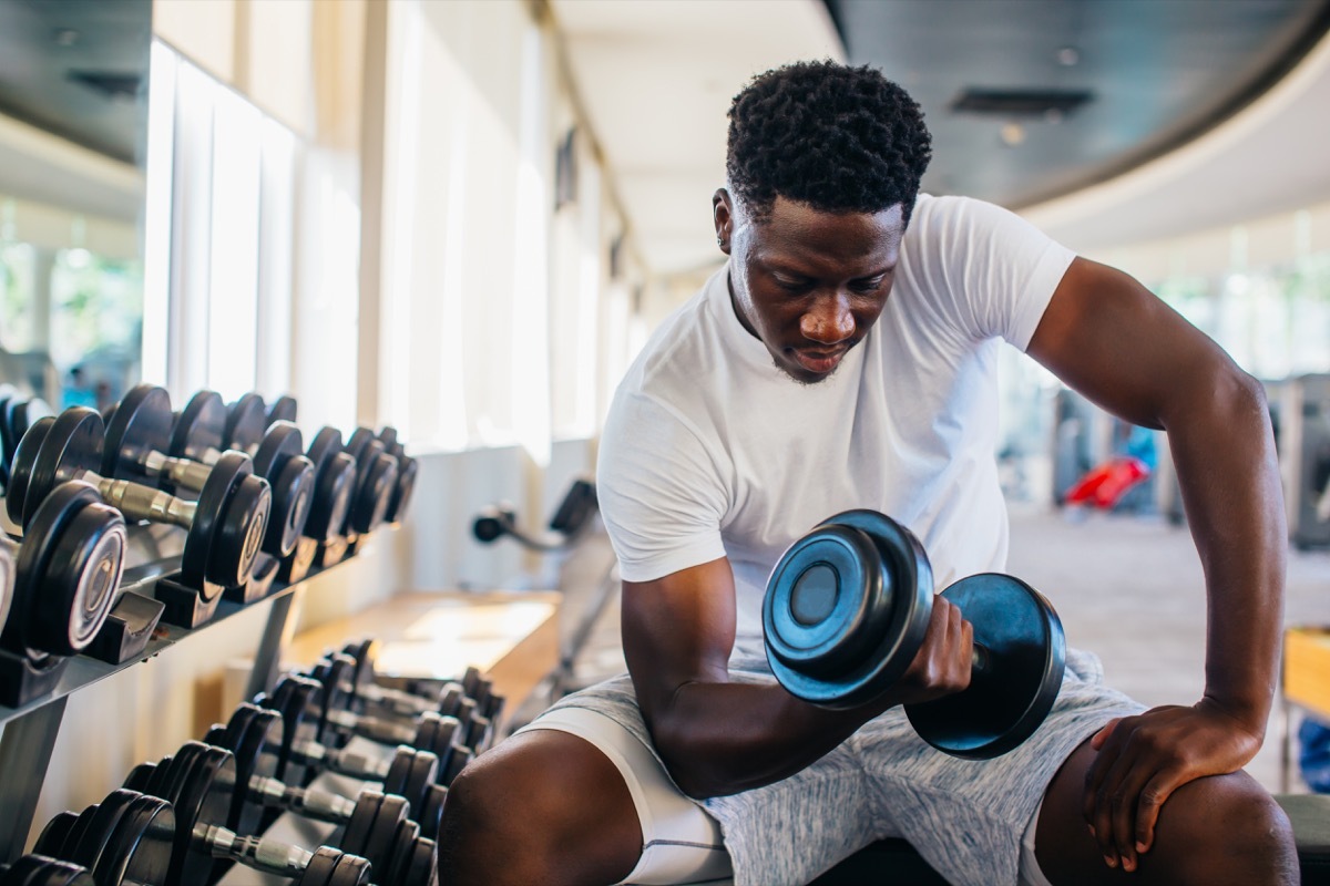 Young man sitting and lifting a dumbbell close to the rack at gym. Male weight training person doing a biceps curl in fitness center