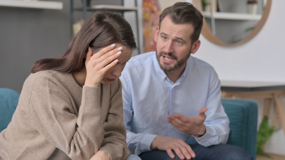 A couple on the couch arguing; the man is talking and the woman has her head in her hands.