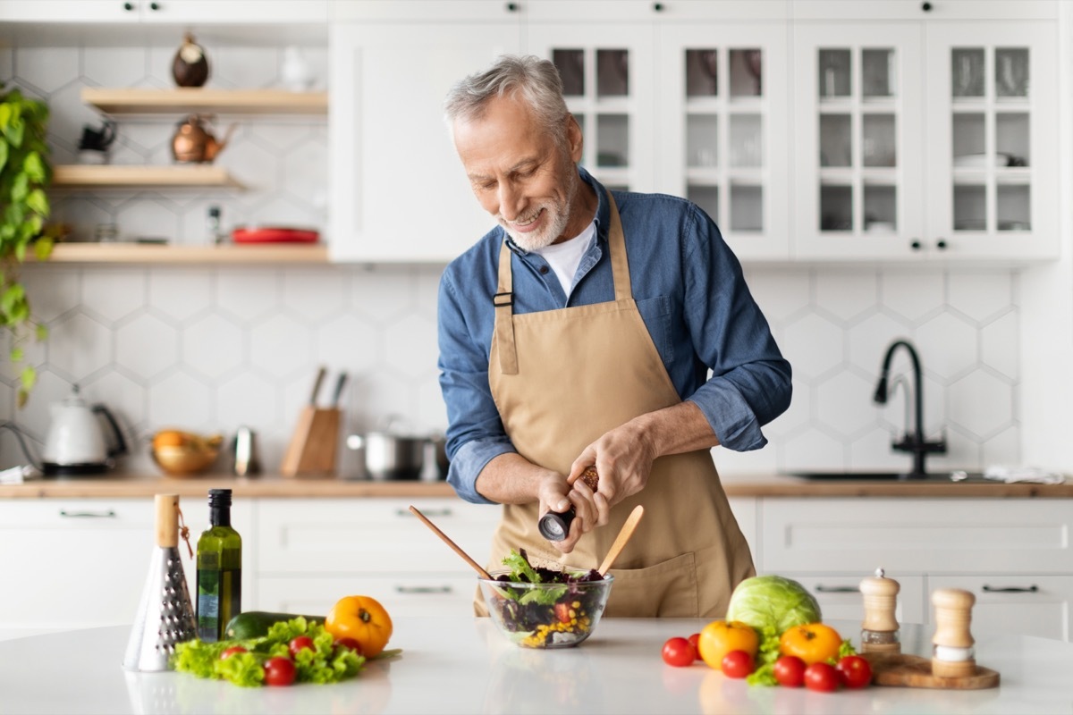 Happy senior man preparing healthy vegetable salad in kitchen, seasoning meal, smiling elderly gentleman in apron adding salt to bowl, enjoying cooking vegetarian food at home, free space