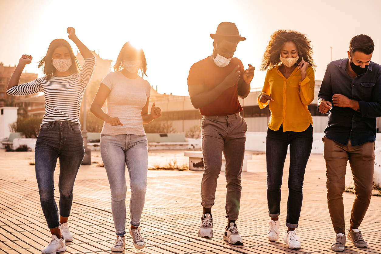 A group of young people wearing face masks dance and celebrate outdoors near sunset.