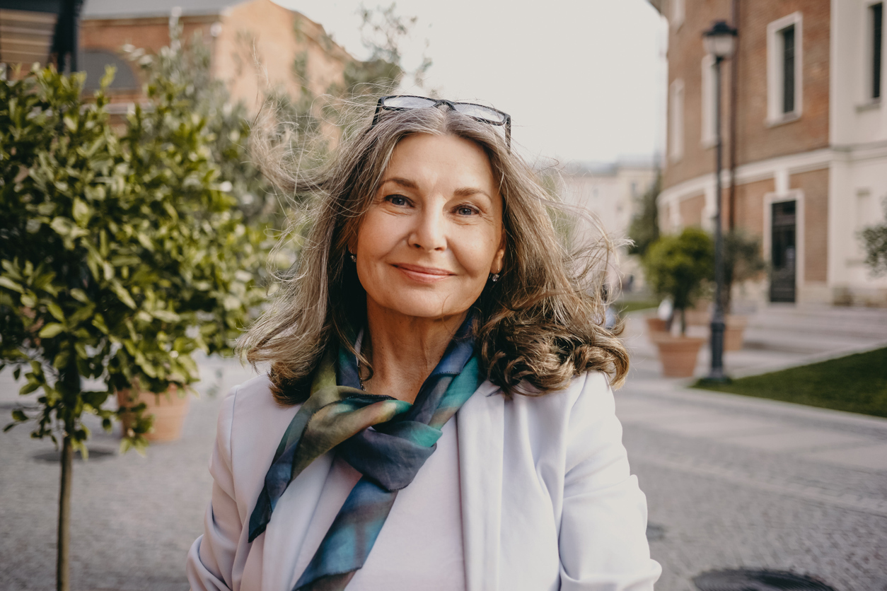 Outdoor image of gorgeous positive lady with charming smile and loose gray hair enjoying nice summer day, wearing white jacket, silk scarf and eyeglasses. Beauty, urban style and fashion concept