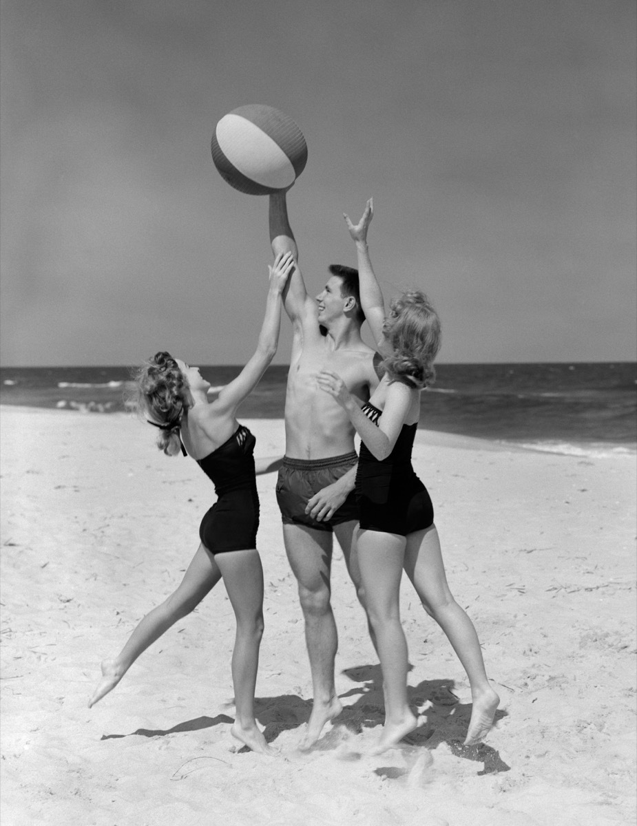 Two 1950s female teens reach for beach ball in male teens hands on beach, cool grandparents