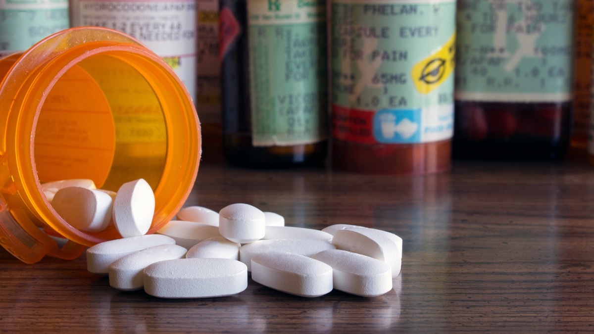 White prescription pills spilled onto a table with many prescription bottles in the background