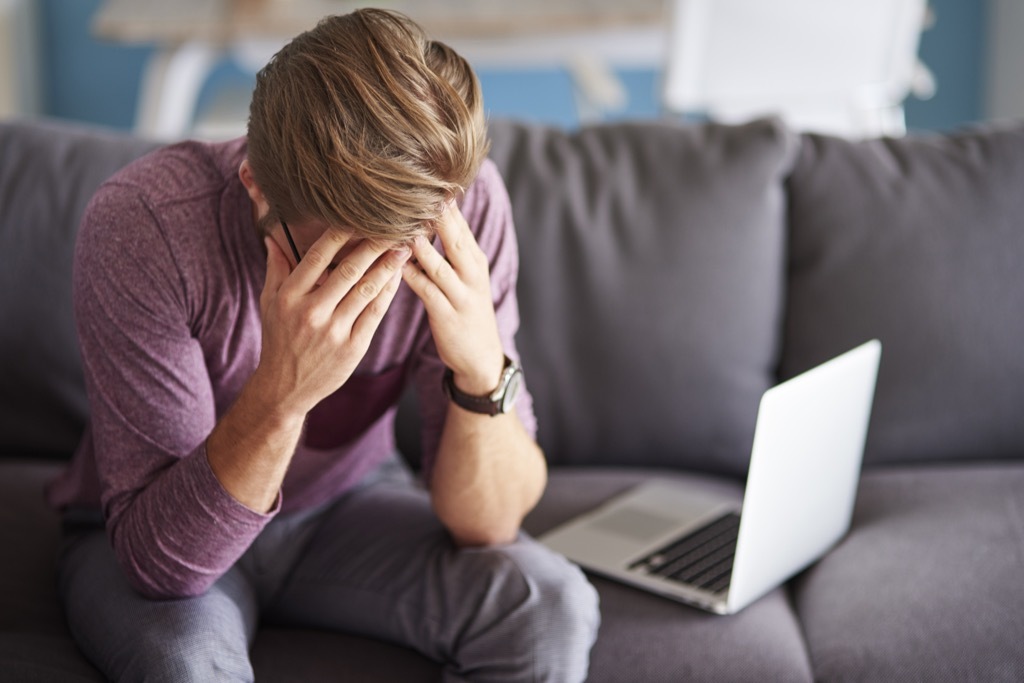 man on couch next to laptop holding head in hands