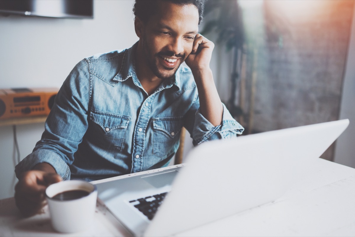 Happy bearded African man making video conversation via modern laptop with partners while holding white cup black coffee at home