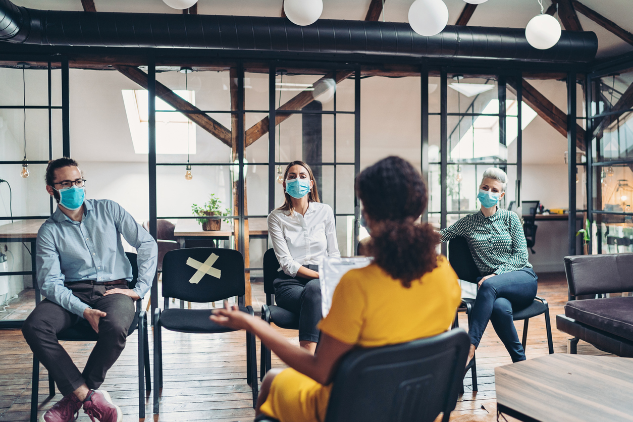 A group of coworkers wearing face masks sit in an office and have a meeting while social distancing.