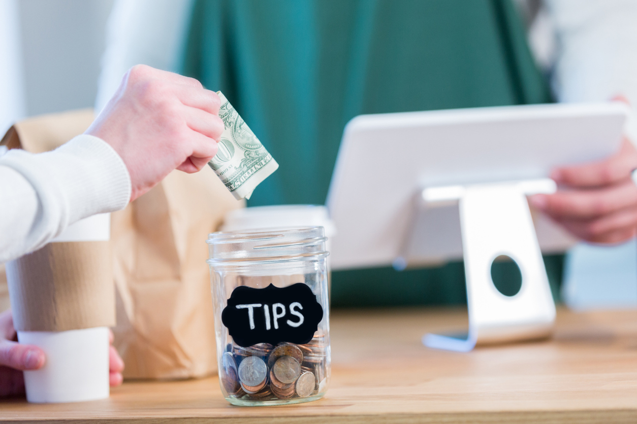 Close up of a someone putting a dollar into a tip jar next to a cash register