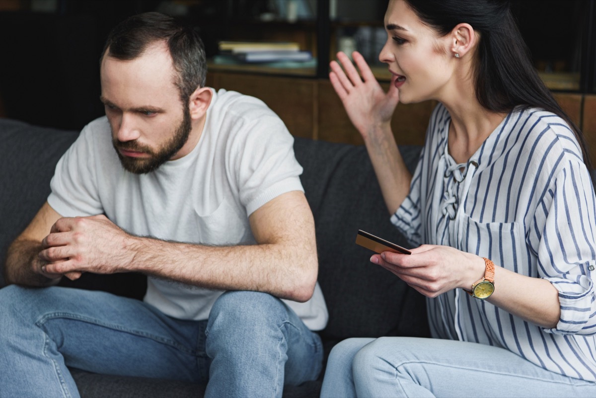 depressed young man sitting on couch and looking away while his wife shouting at him and holding credit card