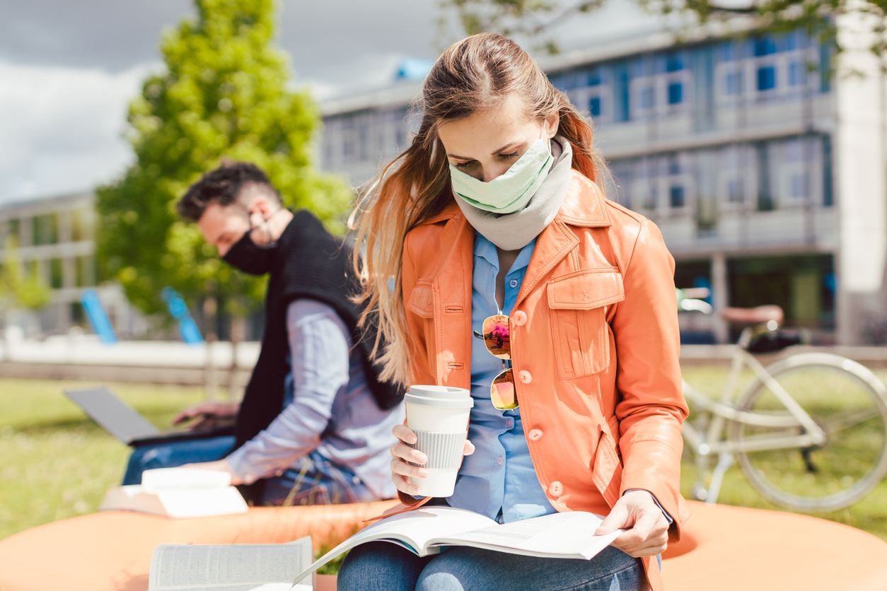 A young female student studies her notes while drinking coffee and wearing a face mask on a college campus with a male student in the background.