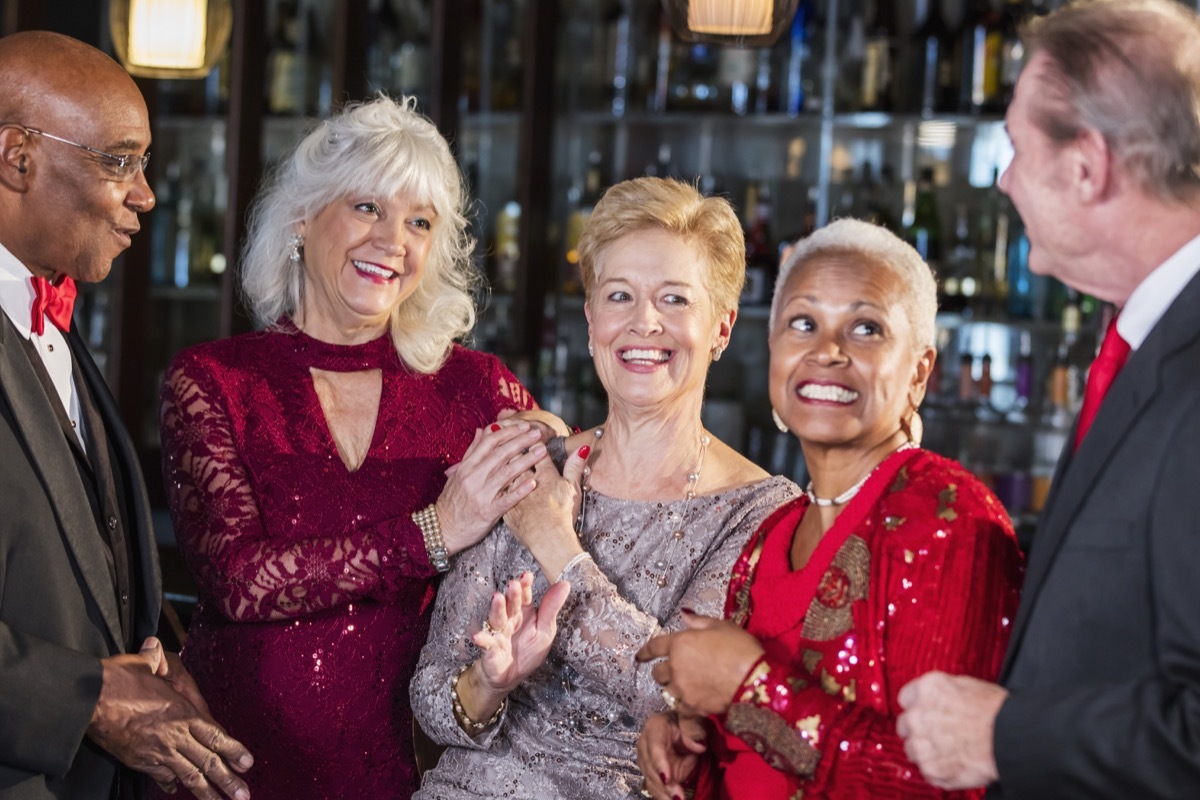 a multi-ethnic group of five seniors enjoying a night out, hanging out together at the bar counter of a restaurant, talking and laughing. They are well-dressed, wearing suits and dresses. The woman in the middle is in her 70s and her friends are in their 60s.