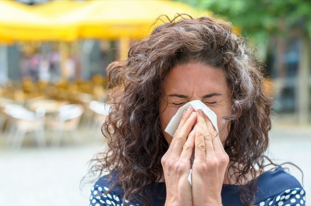 woman with lovely long curly hair standing outdoors blowing her nose in an urban square due to a seasonal cold or hay fever