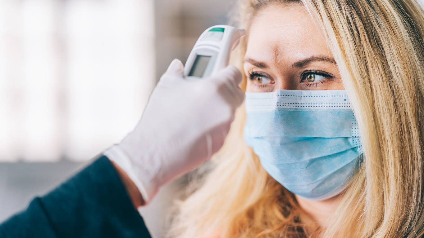 A blonde woman wearing a mask has her temperature taken on her forehead by a health care worker.