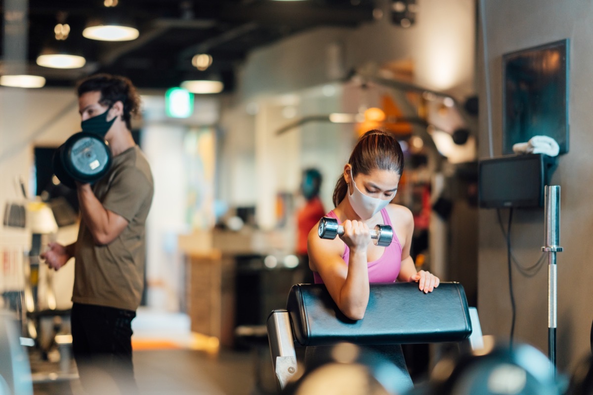 Two sports persons are wearing protective face masks and training in a gym while keeping social distancing.