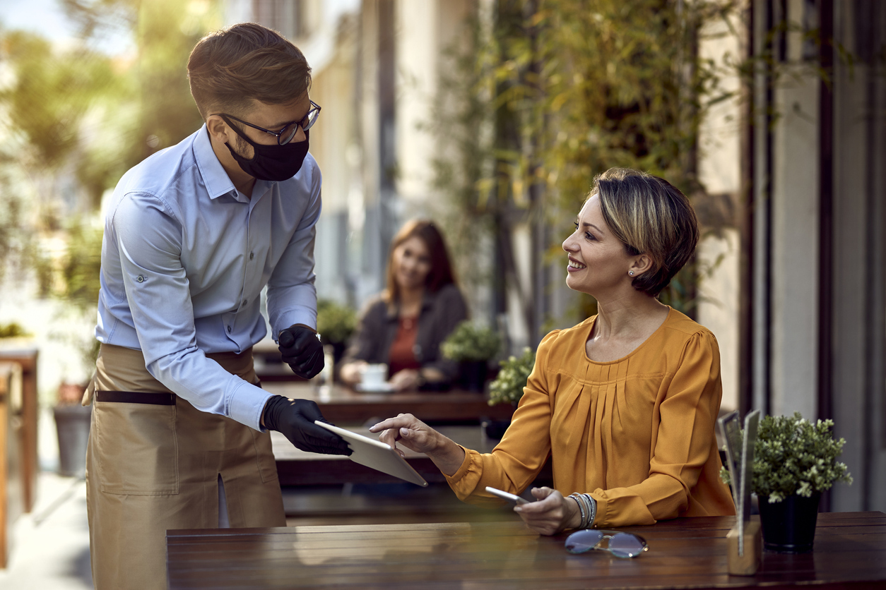 A woman seated at a table without a face mask orders from a waiter wearing PPE