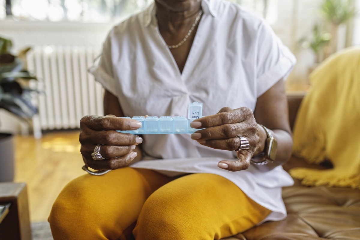 oman sits on the couch at home and takes medications from a daily pill organizer. Cropped shot does not show the woman's face.