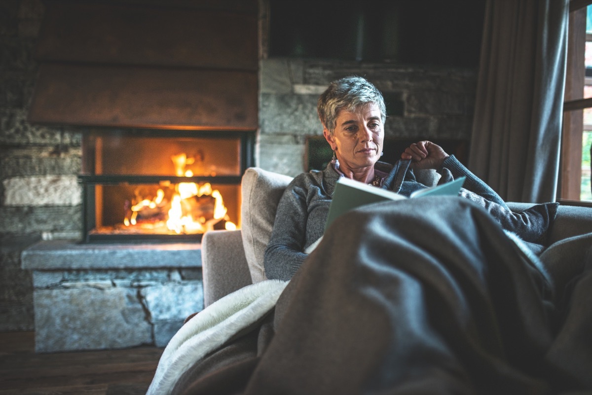 older white woman reading a book under a blanket by the fire