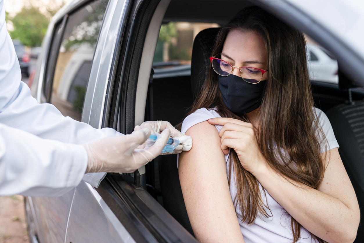 A young woman sitting in a car and wearing a face mask receives the COVID-19 vaccine from a healthcare worker wearing gloves.