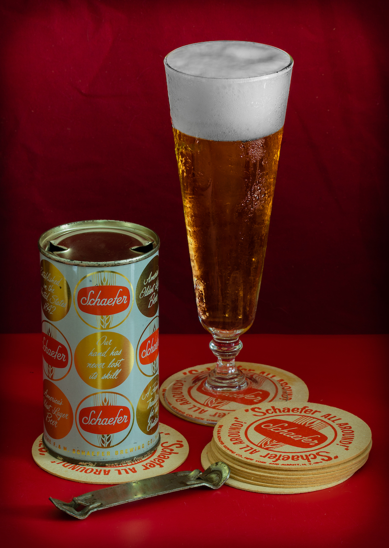 Flat top Schaefer beer can with a pilsner glass of beer, can opener and Schaefer all around coasters on a red table and red background