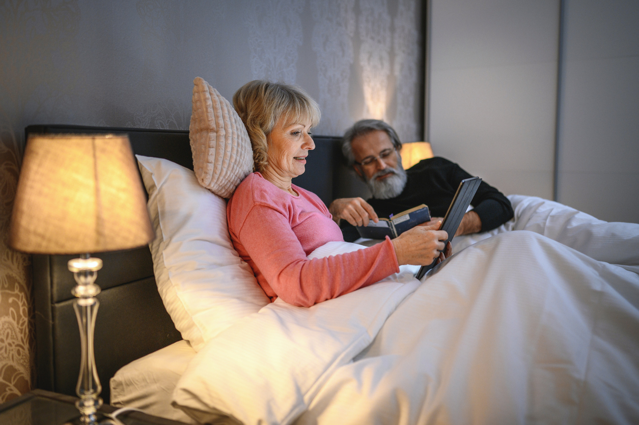 A senior couple lying in bed while using a tablet and reading before going to sleep