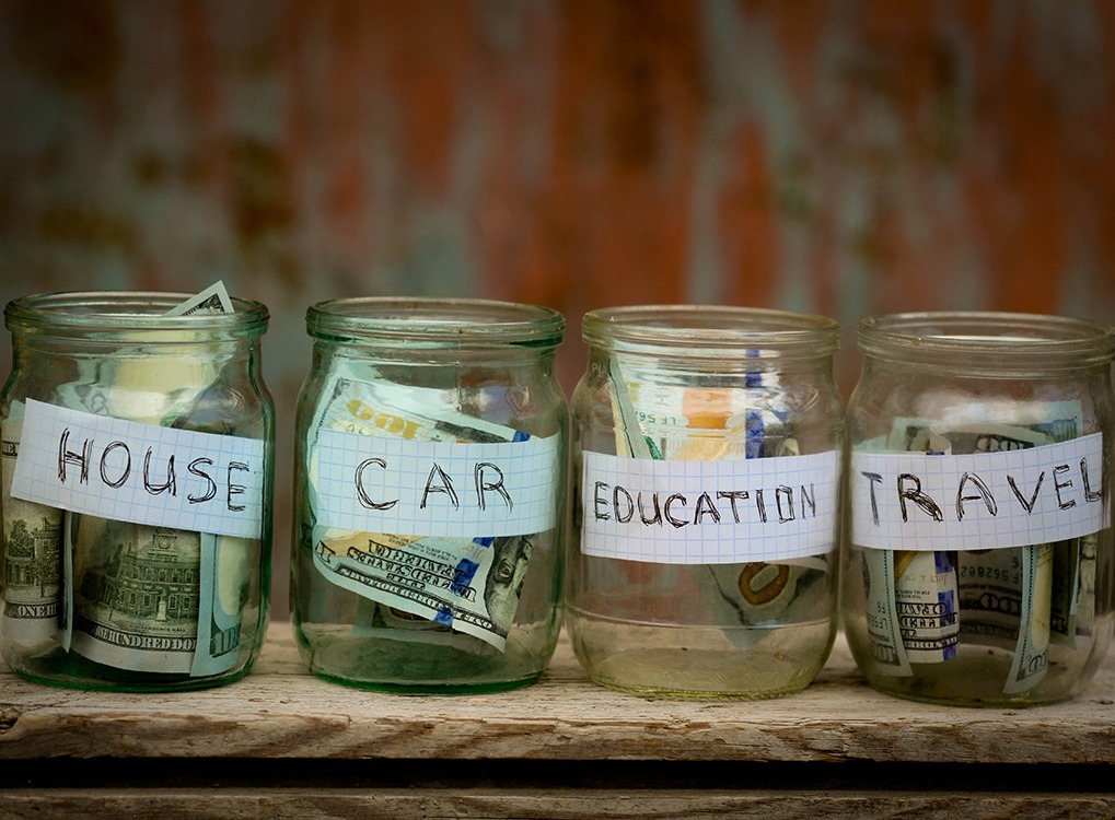 four clear jars labeled house, car, education, and travel, stuffed with dollars