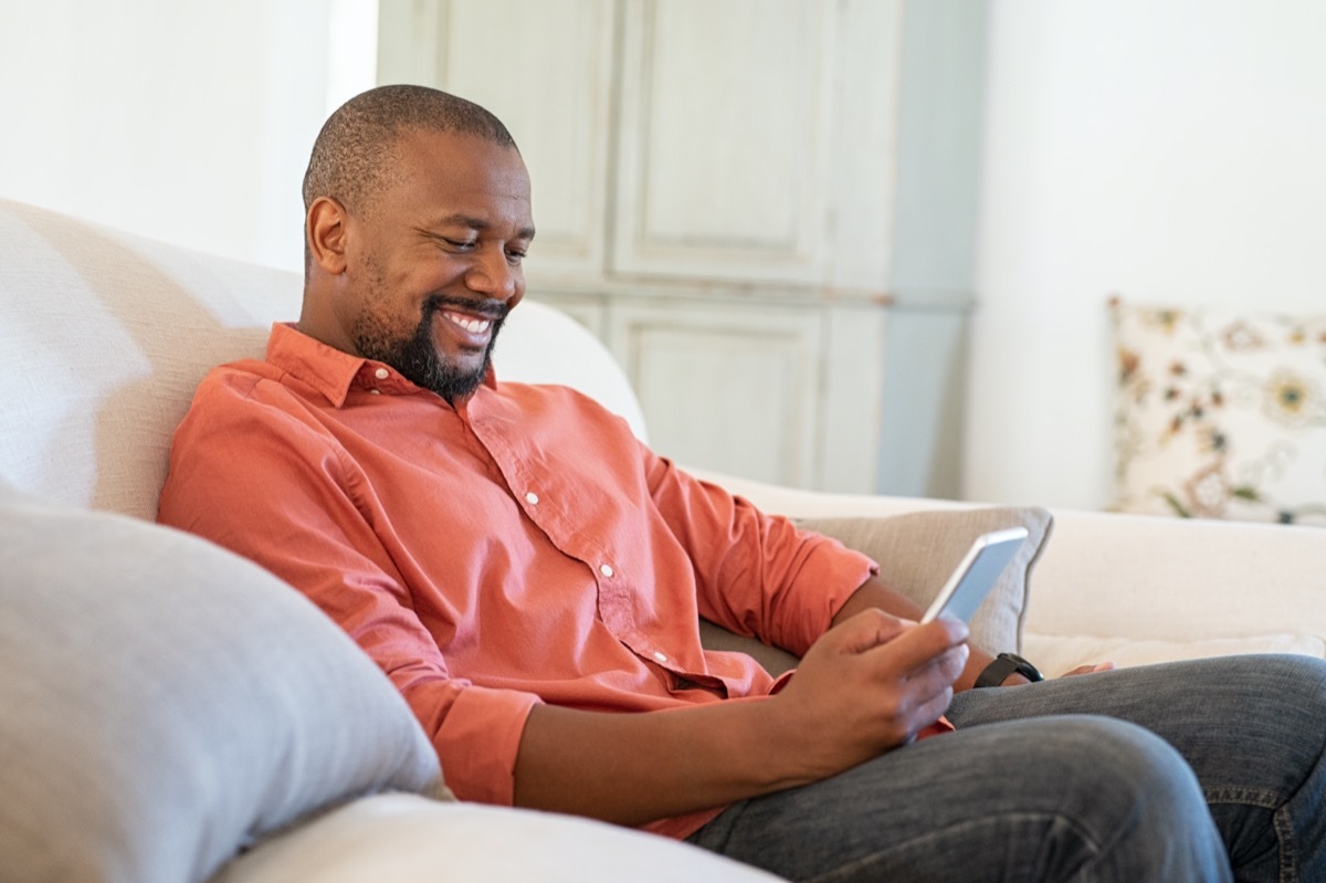 man using smart phone while relaxing at home. Smiling mature man at home sitting on couch reading phone message.