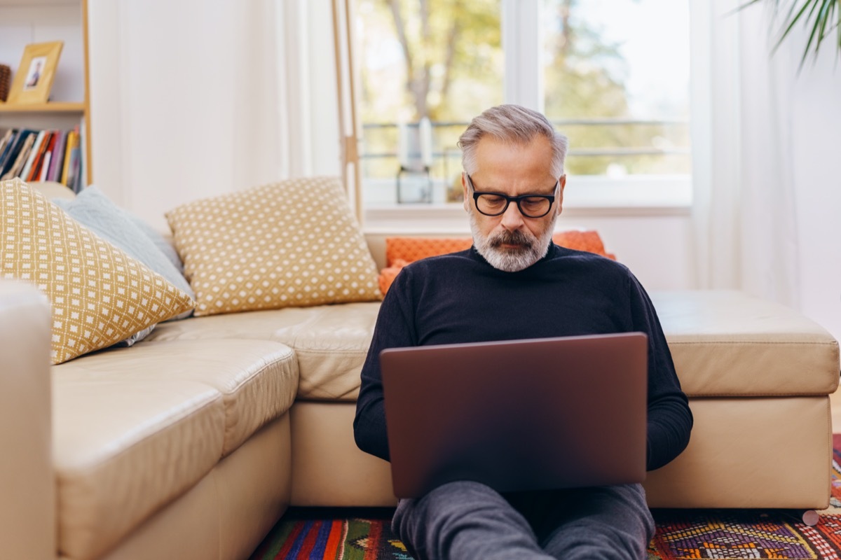 Senior man sitting using a laptop at home relaxing on the floor of the living room leaning against the sofa