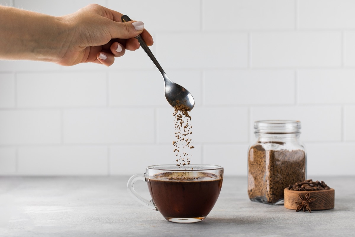 Woman stirs instant coffee in glass mug with boiled water on grey stone table