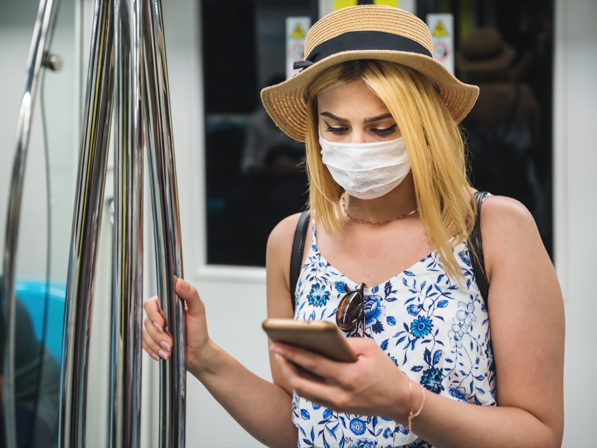 woman wears face mask and keeps social distance by standing and using smart phone while transporting at train