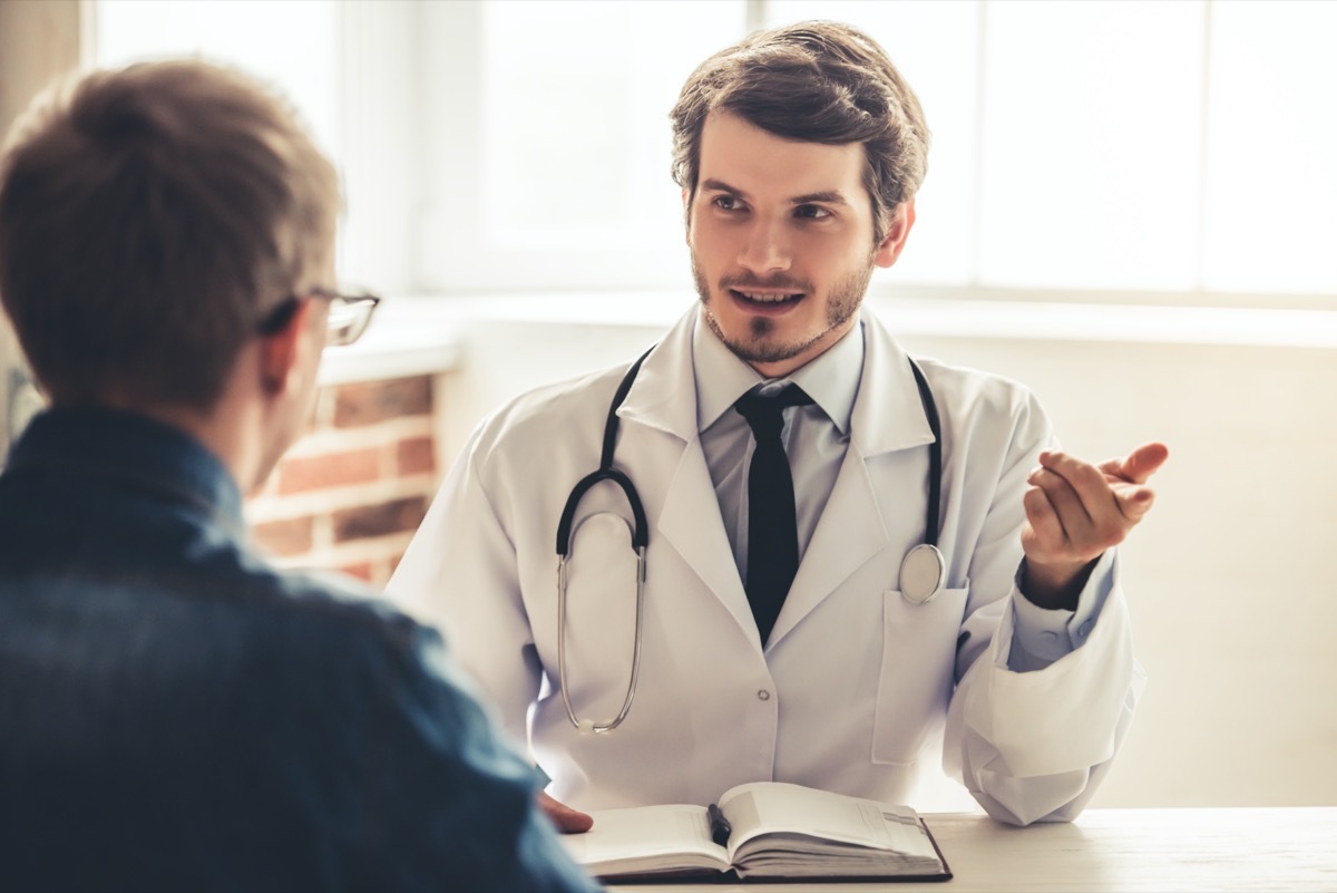 Handsome young doctor in white coat is talking to his patient while working in office