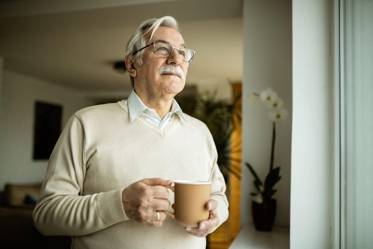 A senior man drinking a cup of coffee or tea from a mug while looking out the window