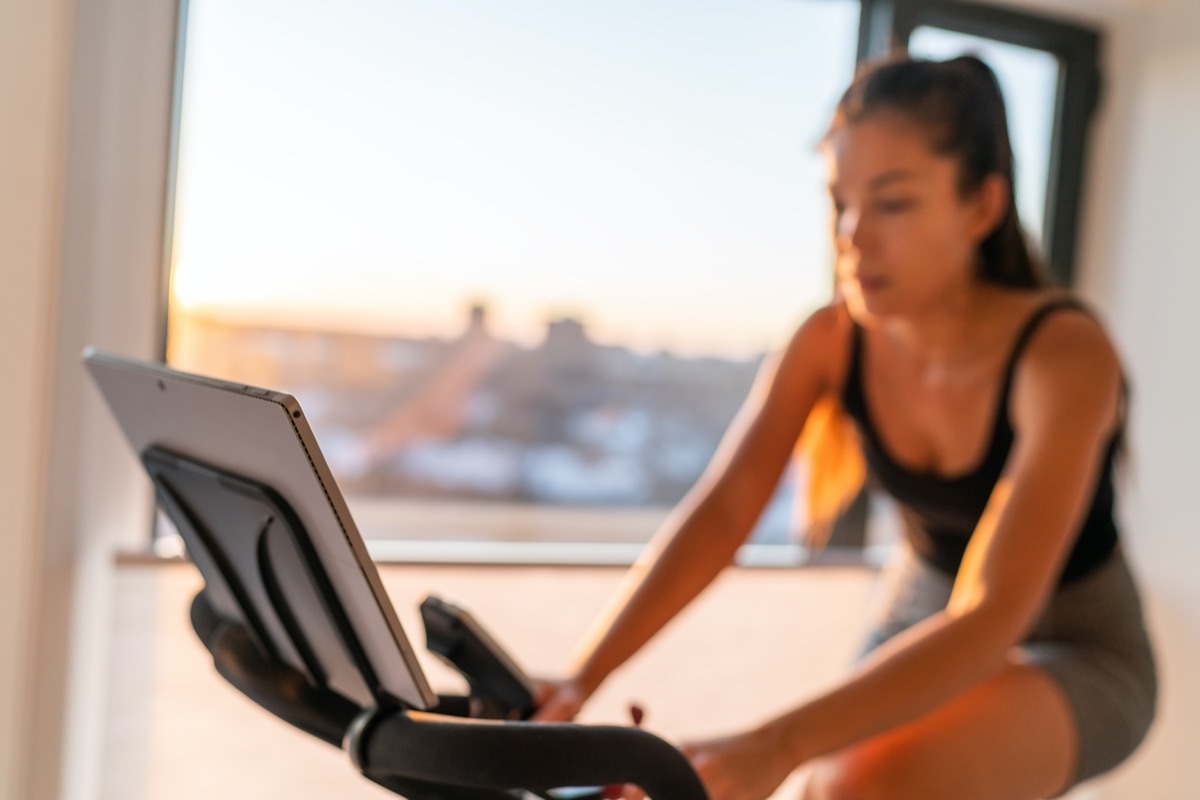 Home fitness fit woman exercising on smart stationary bike at home gym class watching screen online class biking exercise