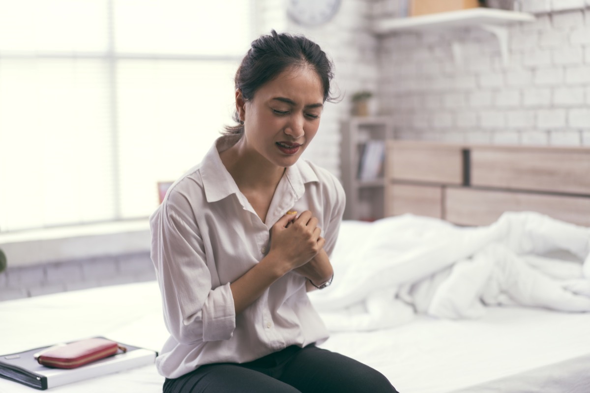 Woman sitting on unmade bed, grimacing and clutching her chest