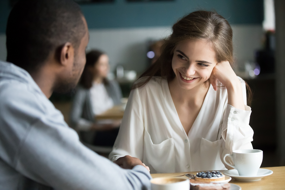 Happy interracial couple flirting talking sitting at cafe table, african man holding hand of smiling caucasian woman having fun drinking coffee together at meeting