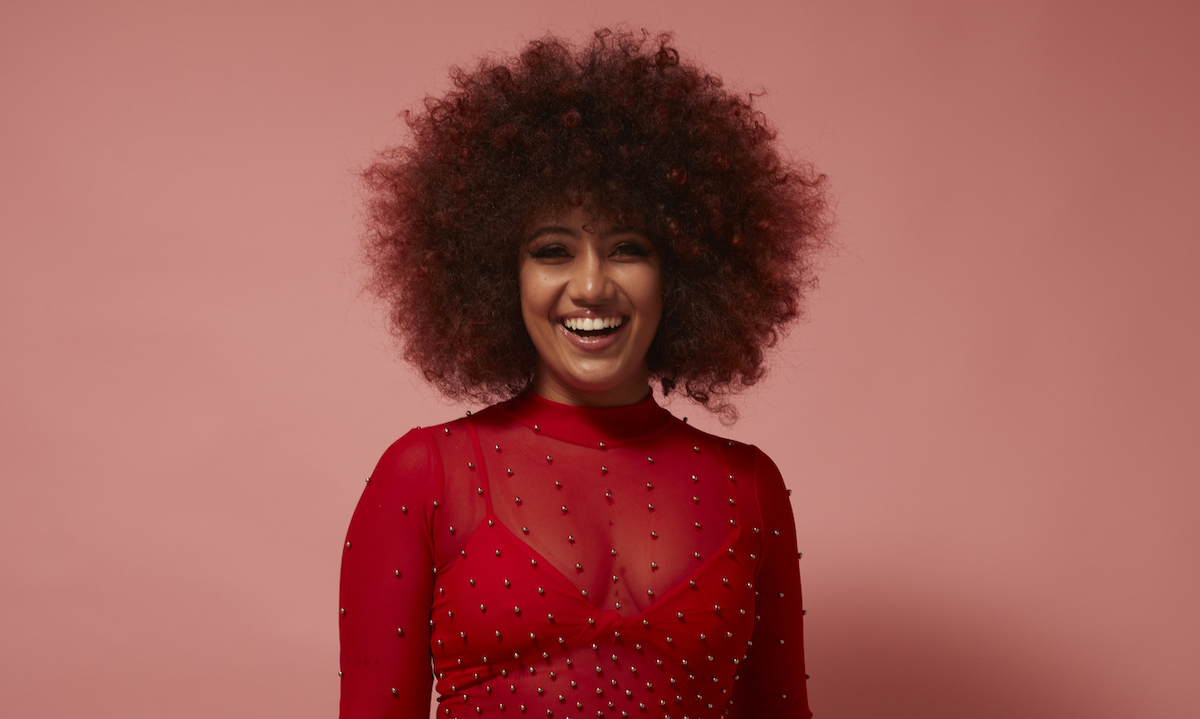 Woman with an afro wearing a sheer red top against a pink background.