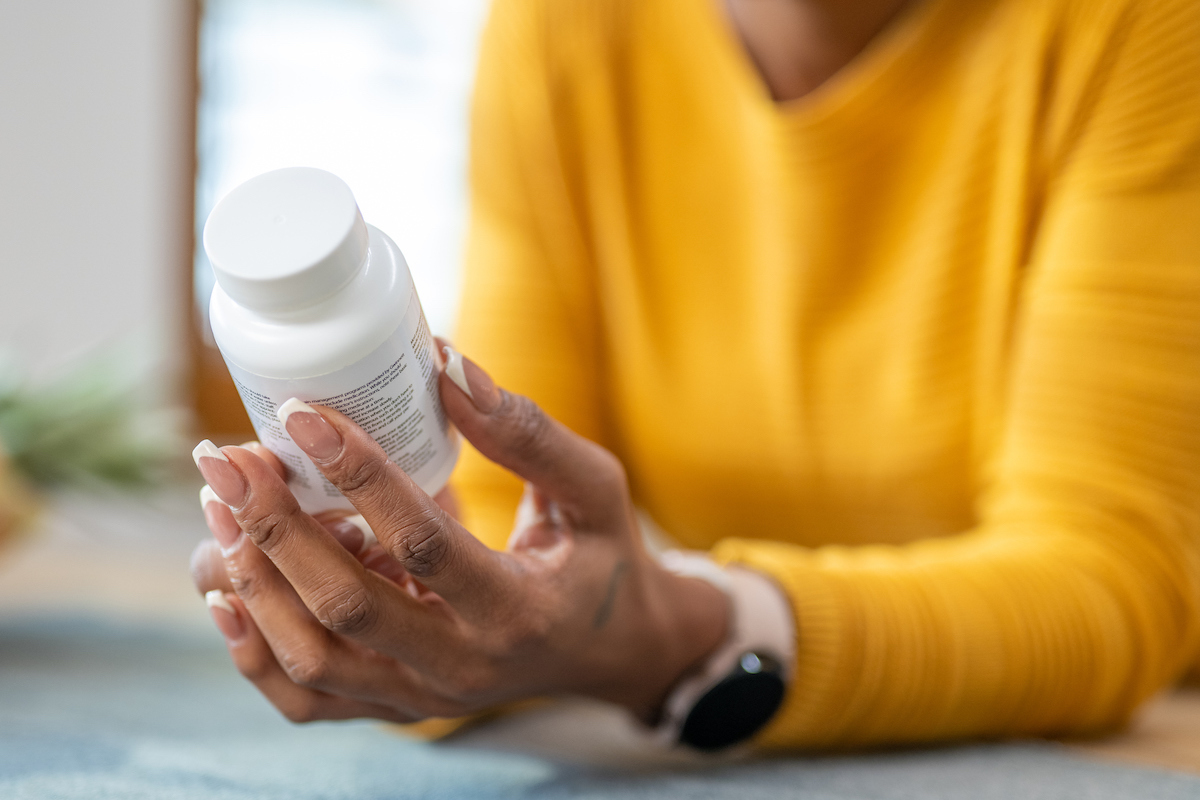 Close up of a woman in a yellow sweater holding a pill, vitamin, or supplement bottle, reading the ingredients