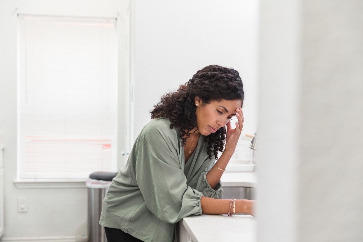 The young adult woman with a severe headache leans against the kitchen counter and rests her head on her hand.