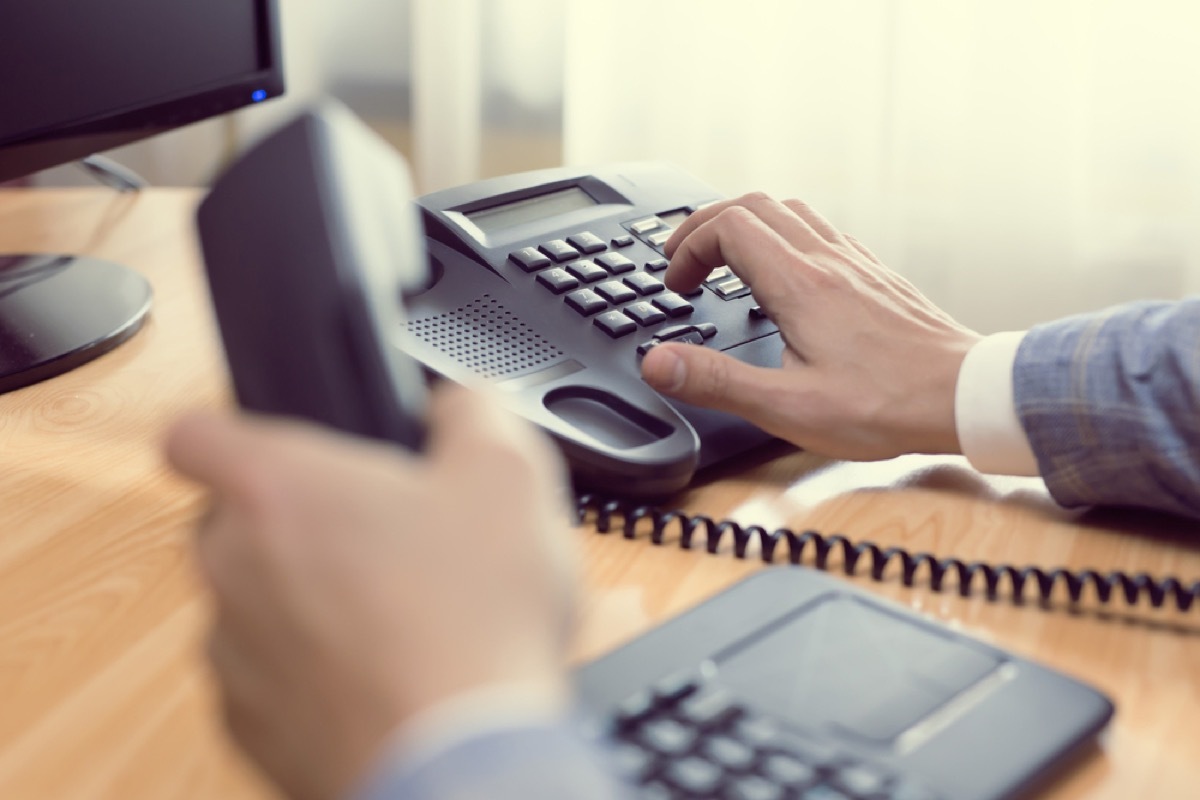 man's hand dials phone while sitting at desk with receiver in other hand, trying to reach a customer service rep