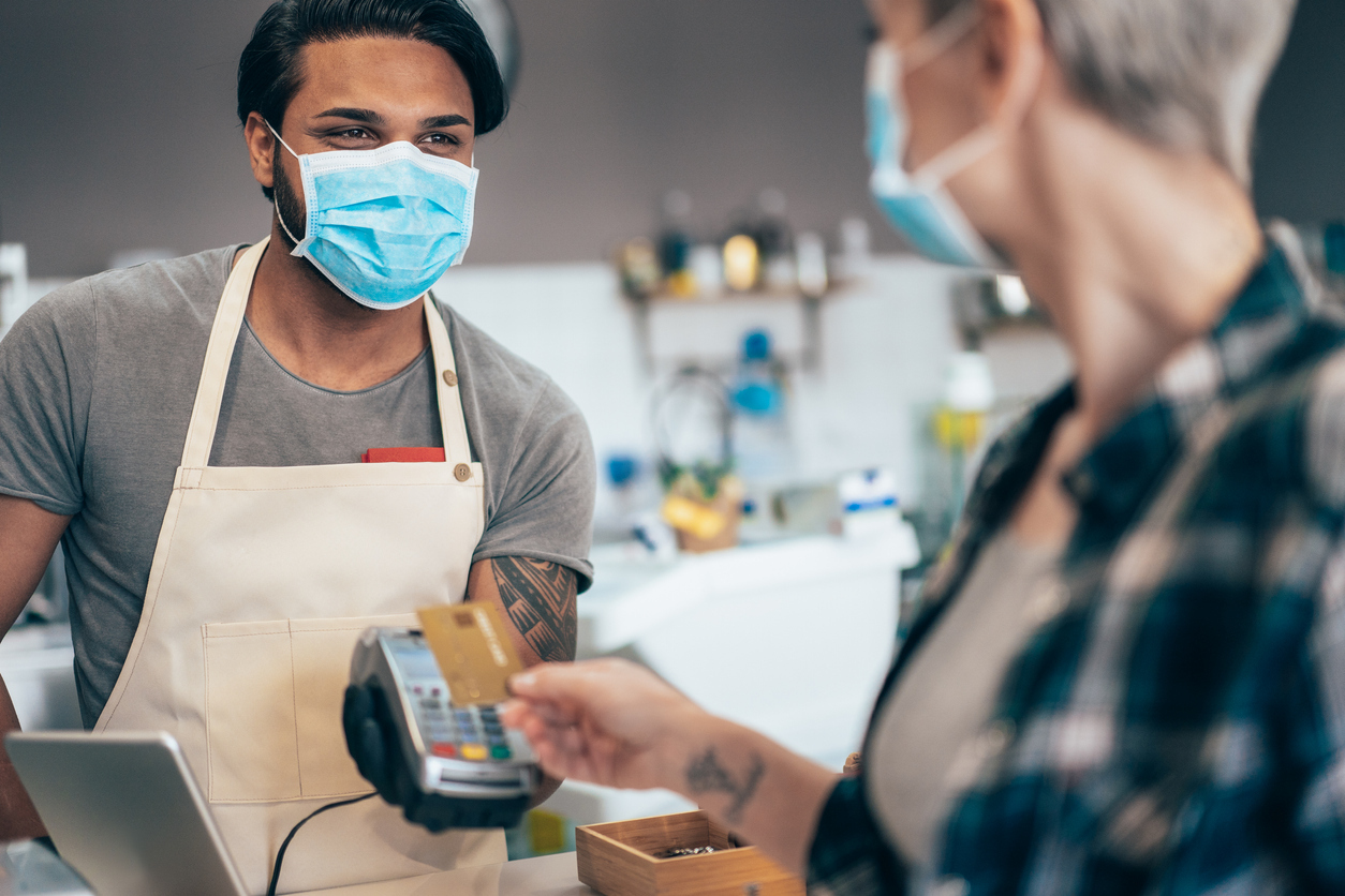 A woman wearing a face mask pays for a coffee with a credit card from a young male barista who is also wearing a face mask and smiling.