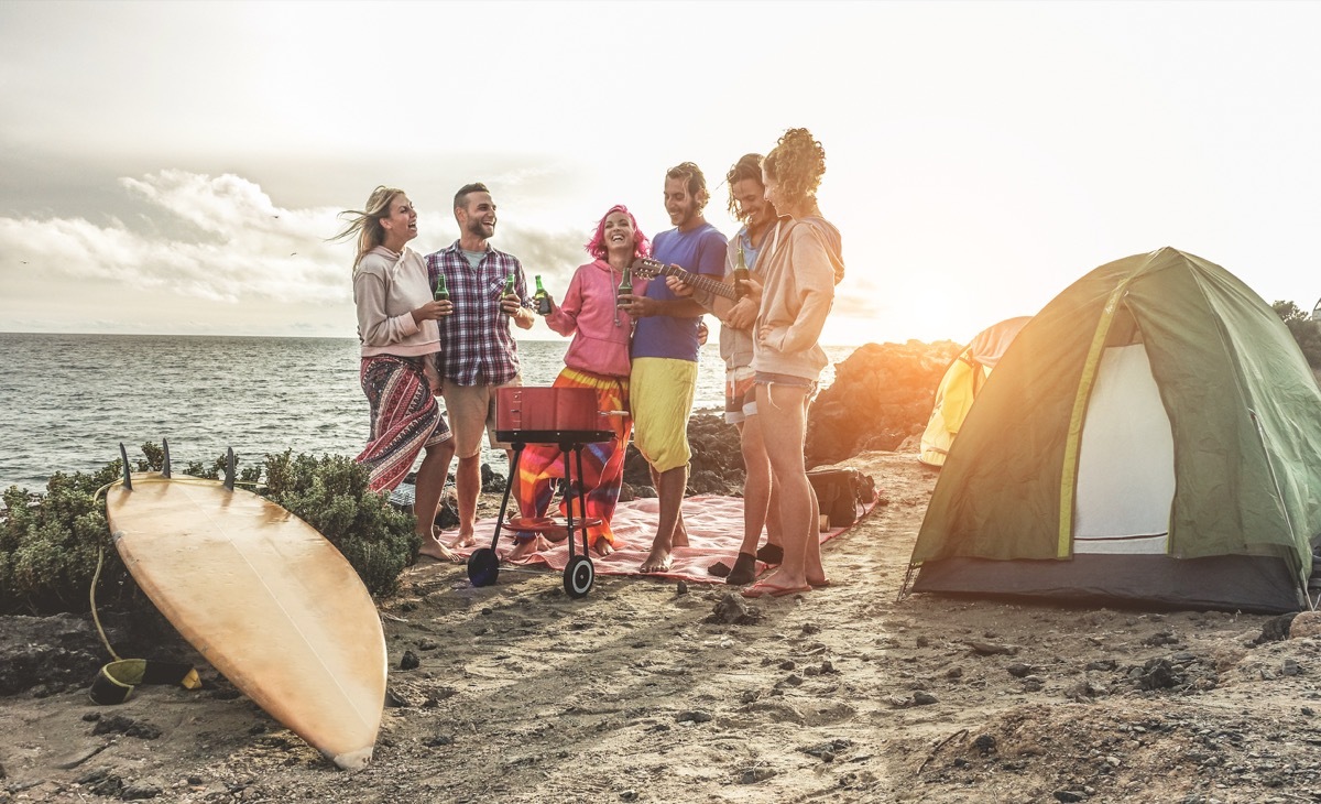 Happy friends drinking beers at camping barbecue picnic next to the ocean - Surfers people having fun and laughing together - Main focus on right guys - Travel, vacation and friendship concept