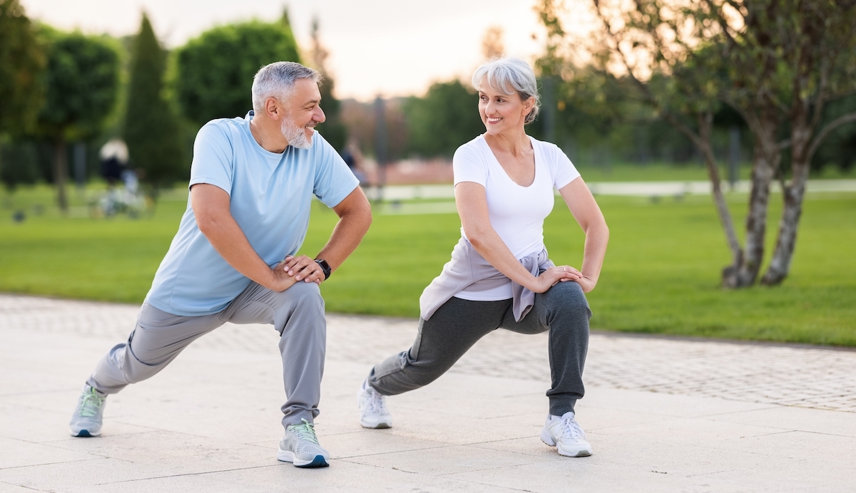 A middle-aged man and woman, both with gray hair, exercise outside. They're stretching doing lunges.