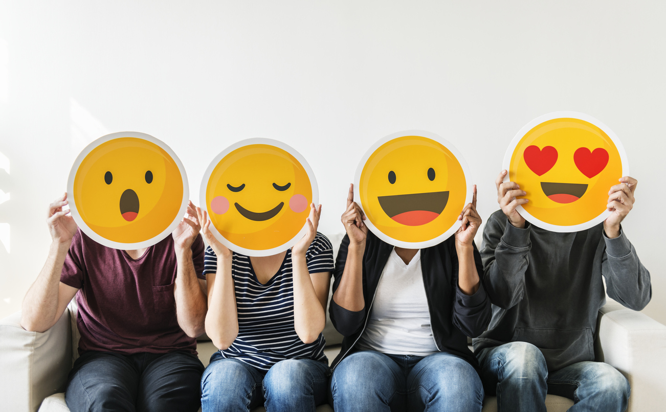 Four people sitting on a couch each holding a different reaction emoji cut out in front of their faces.