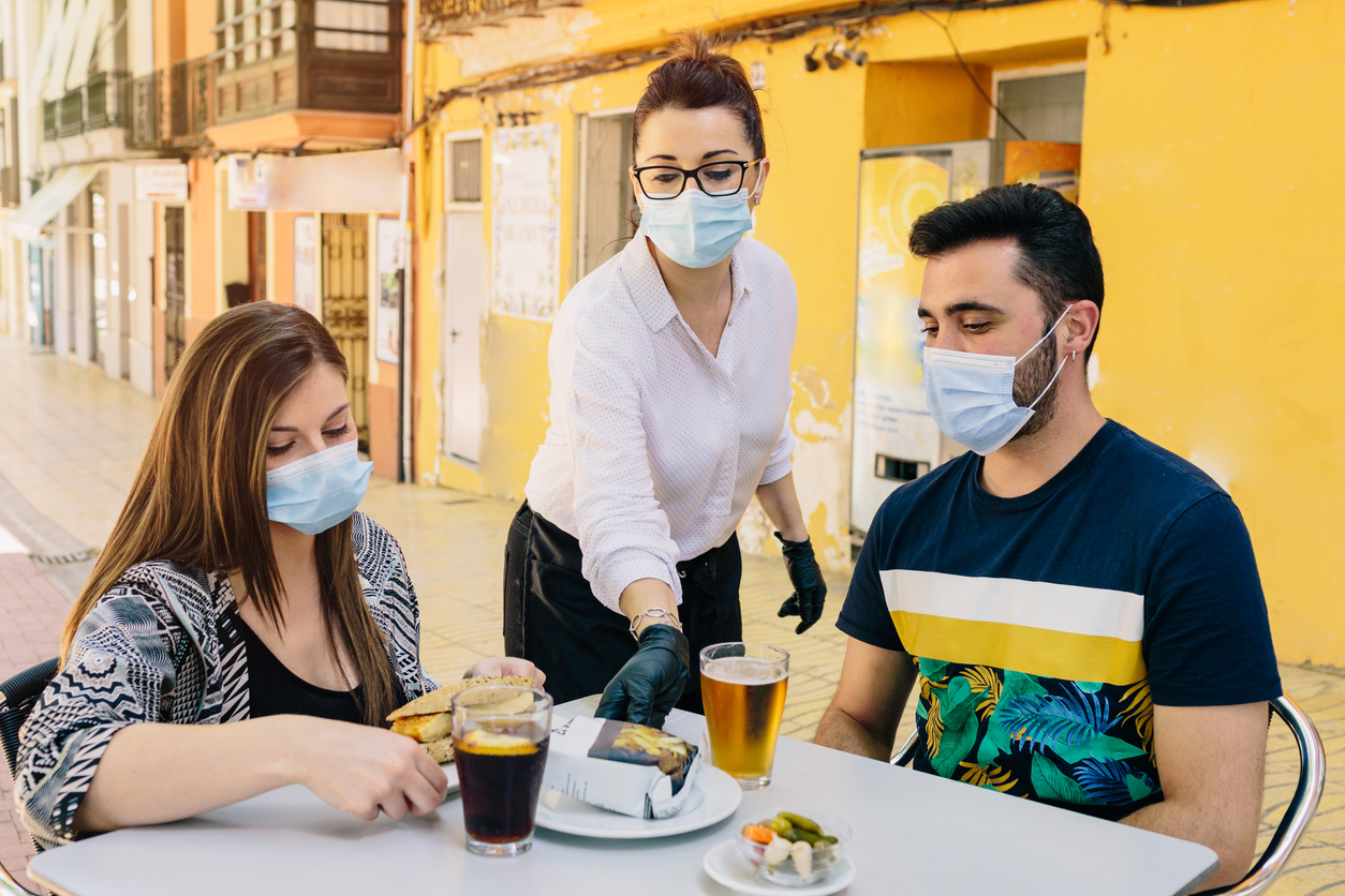 A male and female couple are waited on by a female server at an outdoor table while all wearing face masks