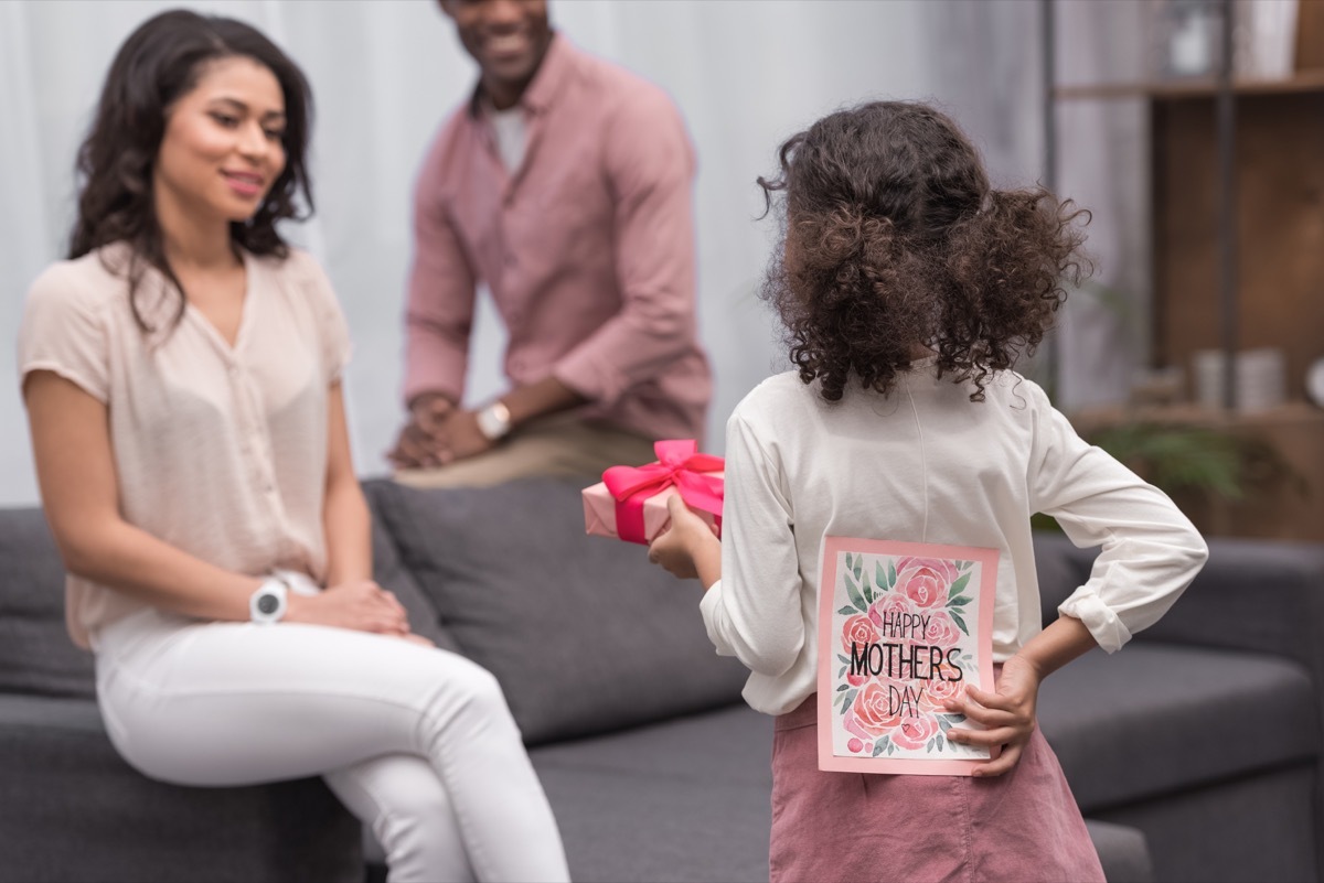 little black girl approaches mother sitting on grey couch with a gift and a happy mother's day card behind her back, state fact about west virginia