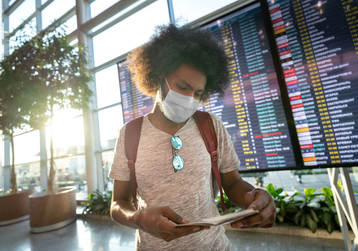 Portrait of a male traveler wearing a face mask at the airport with the flight schedule at the background while looking at his boarding pass - travel concepts 