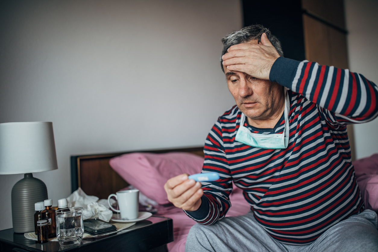 A man with a fever grabs his head with his hand while reading a thermometer, doing a temperature check for coronavirus