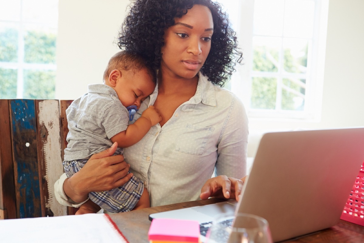 black woman holding sleeping baby while typing on laptop