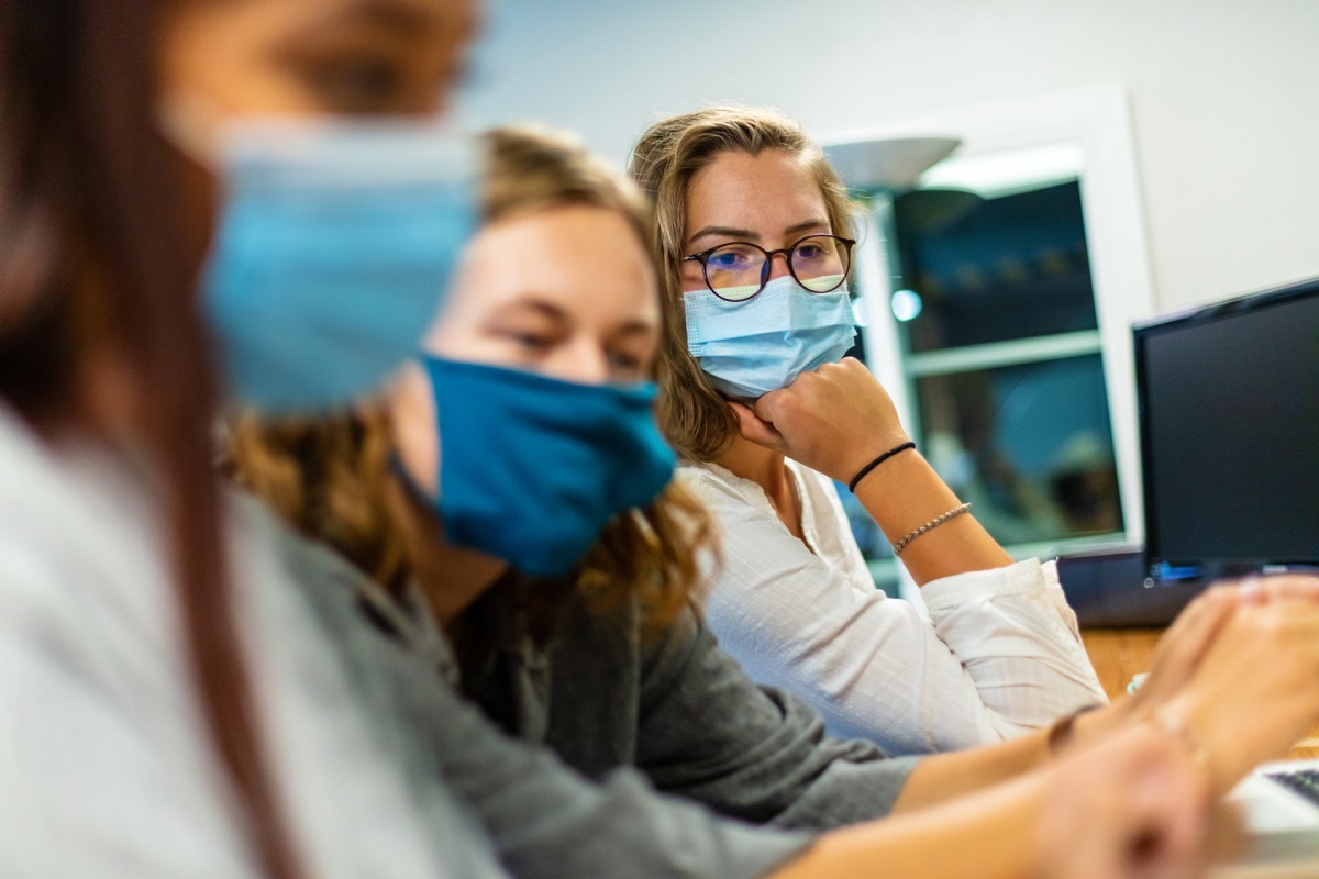 Three female college students; Two in foreground blurred; one in background in focus; wearing face masks using technology working on laptops part of a series (Shot with Canon 5DS 50.6mp photos professionally retouched - Lightroom / Photoshop - original size 5792 x 8688 downsampled as needed for clarity and select focus used for dramatic effect)