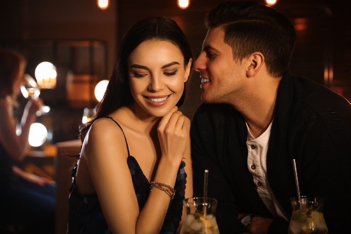 woman smiling seductively while man whispers in her ear at a bar