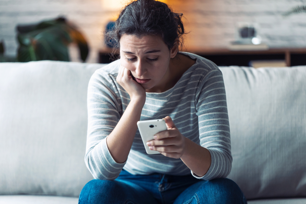 A young woman sitting on a couch and looking at her phone with a concerned expression on her face